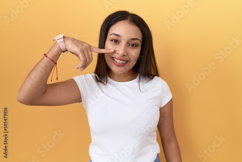 Young arab woman wearing casual white t shirt over yellow background pointing with hand finger to face and nose, smiling cheerful. beauty concept