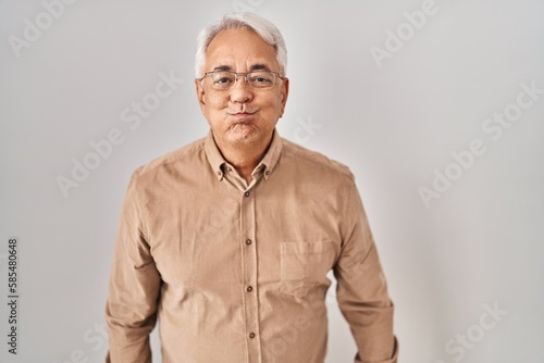 Hispanic senior man wearing glasses puffing cheeks with funny face. mouth inflated with air, crazy expression. © Krakenimages.com