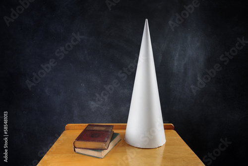 Dunce cap on a desk with books, negative words written on the chalkboard photo