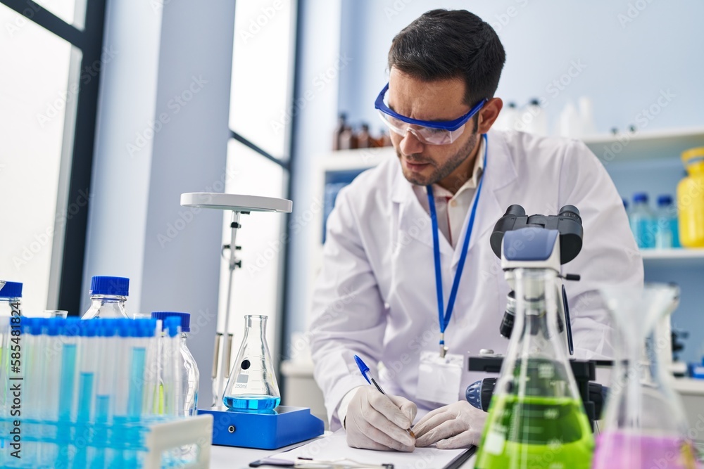 Young hispanic man scientist measuring liquid writing report at laboratory Young hispanic man scientist measuring liquid writing report at laboratory