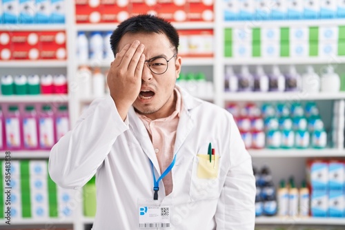 Chinese young man working at pharmacy drugstore yawning tired covering half face, eye and mouth with hand. face hurts in pain. © Krakenimages.com