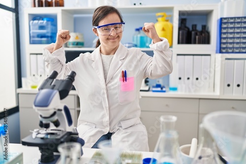 Hispanic girl with down syndrome working at scientist laboratory showing arms muscles smiling proud. fitness concept.