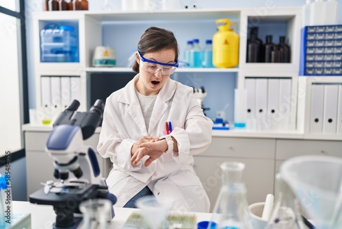 Hispanic girl with down syndrome working at scientist laboratory looking at the watch time worried, afraid of getting late
