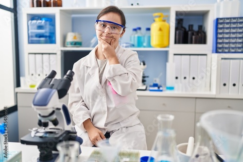 Fototapeta Naklejka Na Ścianę i Meble -  Hispanic girl with down syndrome working at scientist laboratory with hand on chin thinking about question, pensive expression. smiling and thoughtful face. doubt concept.
