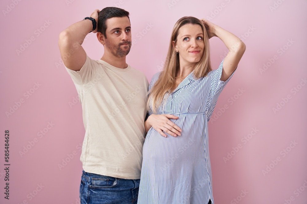 Young couple expecting a baby standing over pink background confuse and wondering about question. uncertain with doubt, thinking with hand on head. pensive concept. Young couple expecting a baby standing over pink background confuse and wondering about question. uncertain with doubt, thinking with hand on head. pensive concept.