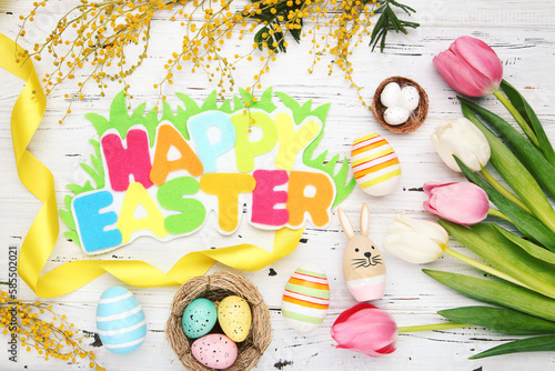 Colorful inscription Happy Easter, eggs in nests, ribbon, flowers of mimosa and tulips on white wooden background