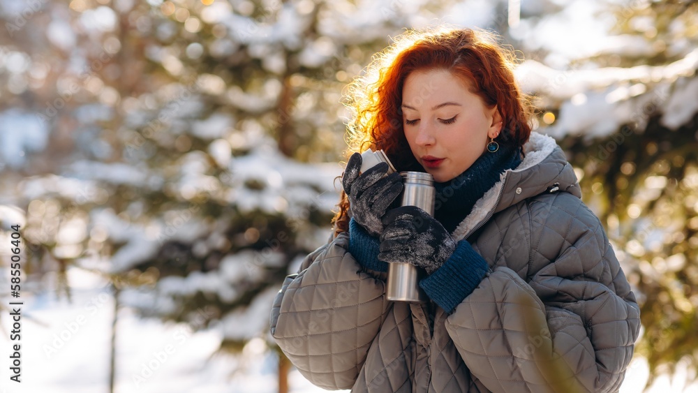 A pretty woman with red hair inhales the smell of fragrant tea from a thermos. A woman in winter clothes stands against the backdrop of snow-covered trees. A pretty woman with red hair inhales the smell of fragrant tea from a thermos. A woman in winter clothes stands against the backdrop of snow-covered trees.