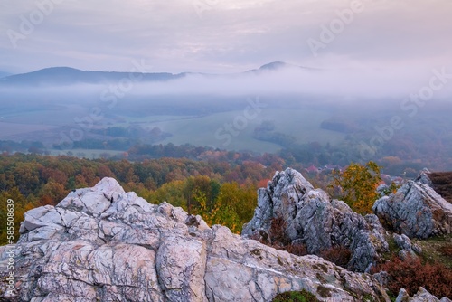 Mountains in low clouds at sunrise in autumn. View of mountain peaks, rocks and colored trees in fog in fall. Beautiful landscape with foggy hills, forest, sunbeam. View from above of mountain valley photo