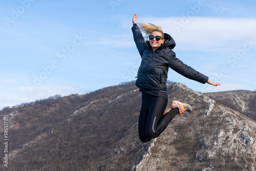 Sporty blonde woman in black dress and sunglasses is jumping on a trip. In the background is a rocky mountainside and blue sky. photo