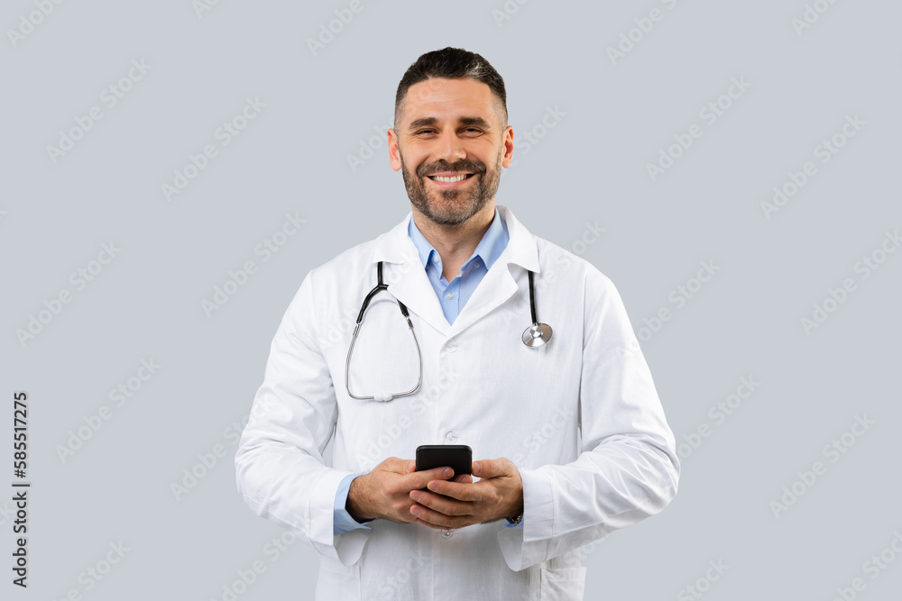 Portrait of friendly male therapist in white coat and with stethoscope using smartphone for work, gray background Portrait of friendly male therapist in white coat and with stethoscope using smartphone for work, gray background