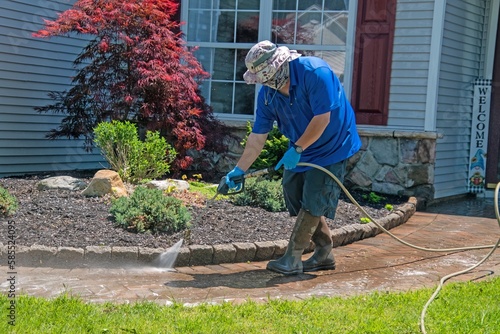 A caucasian man with a latex holding water sprayer wand power washing the brick walkway of house