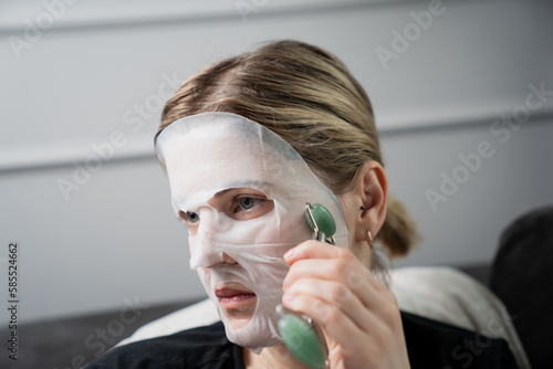Woman using face massage roller on white mask for facial