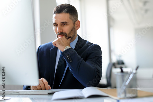 Concentrated middle aged male entrepreneur working with computer, looking at screen and thinking, office interior
