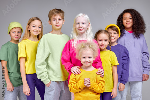 cheerful kids in fashion stylish clothes standing in row behind little girl with fluffy curly fair hair  children support little kid  help her