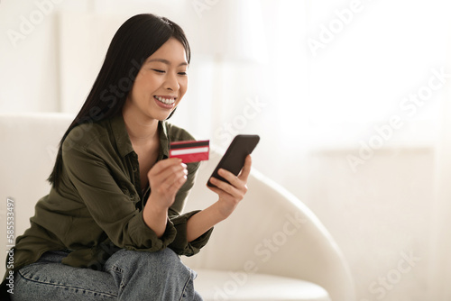 Cheerful asian woman using smartphone and credit card. home interior