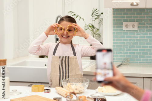 Caucasian young mom takes photo on smartphone of little daughter in apron, makes cookies, puts forms