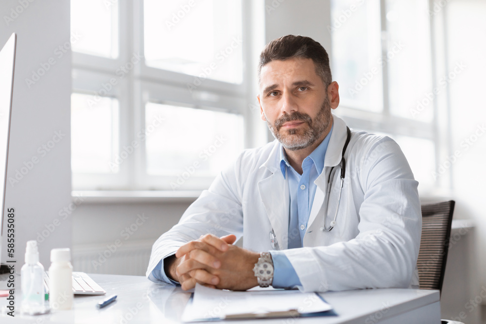 Portrait of confident male middle aged doctor, physician or cardiologist in white coat uniform sitting at desk in office
