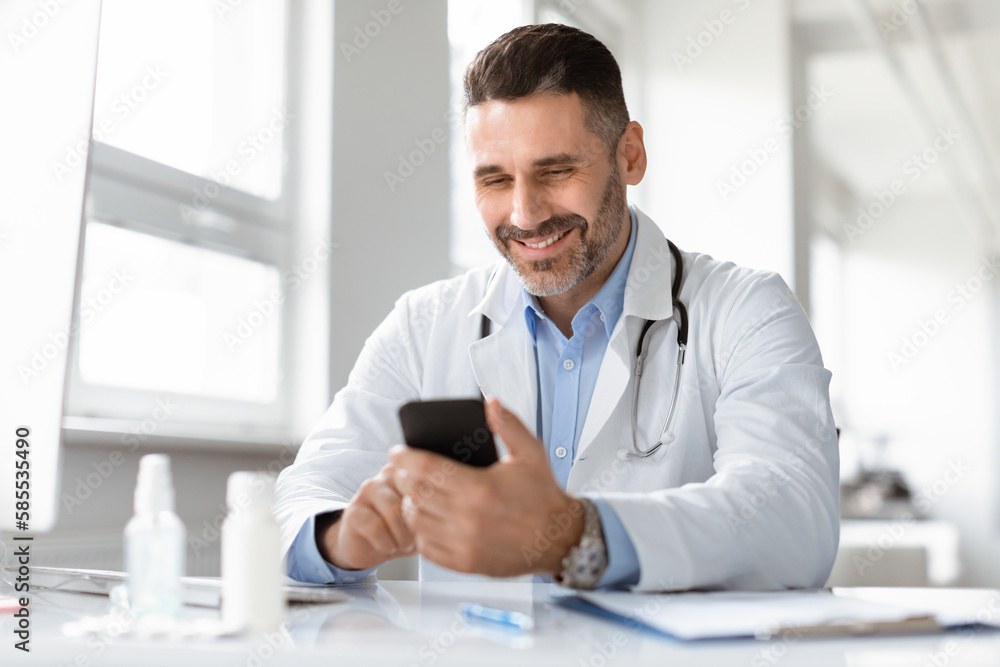 Fototapeta premium Smiling male doctor in medical uniform sitting at desk in clinic, looking at phone screen, consulting client online