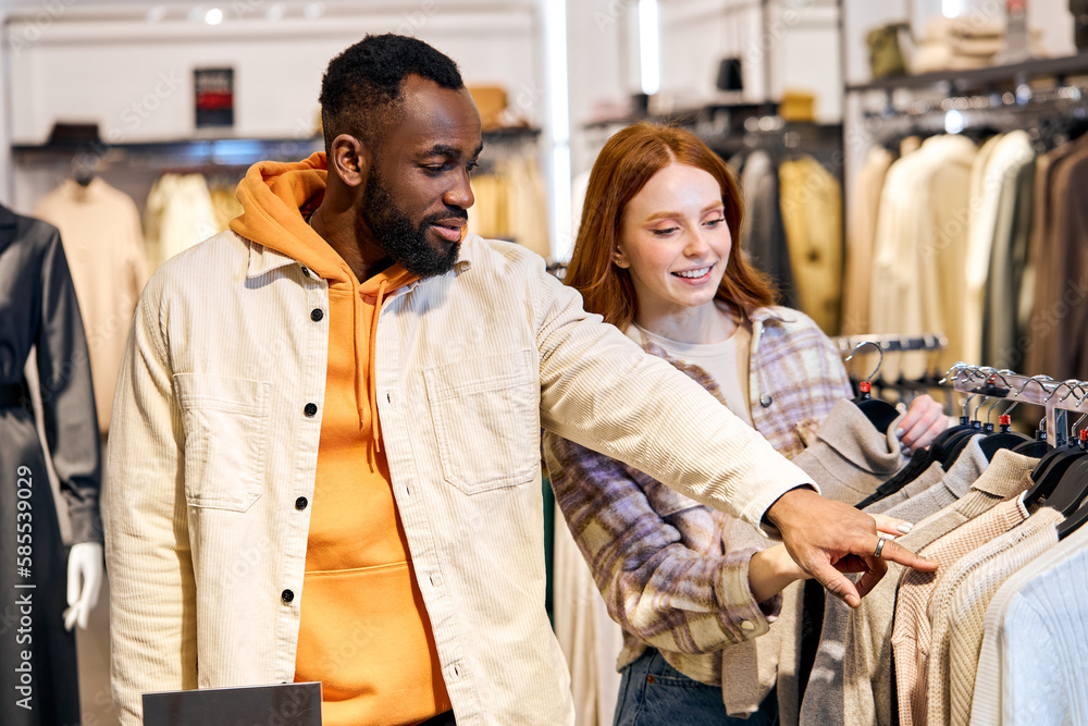 afro handsome guy helps to choose a jacket, sweater for his ginger girlfriend at shop, showroom. close up side view photo, afro handsome guy helps to choose a jacket, sweater for his ginger girlfriend at shop, showroom. close up side view photo,