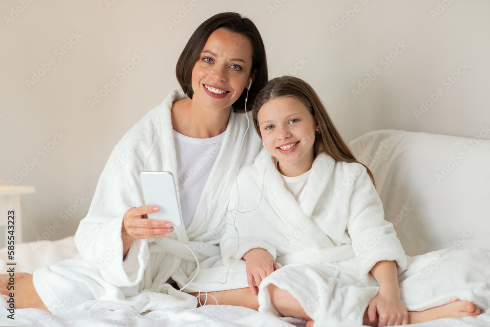 Smiling caucasian small girl, young female in bathrobe sit on white bed with headphones and phone Smiling caucasian small girl, young female in bathrobe sit on white bed with headphones and phone