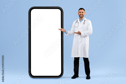 Middle aged male physician showing big empty cellphone screen, posing on blue studio background, full length, mockup