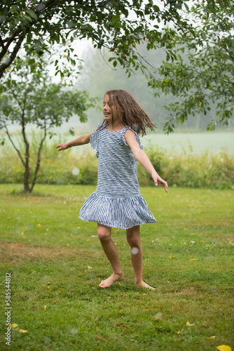 Happy cute little girl in sundress dancing and running on green lawn under the rain. Barefoot child enjoying summer outdoors. 