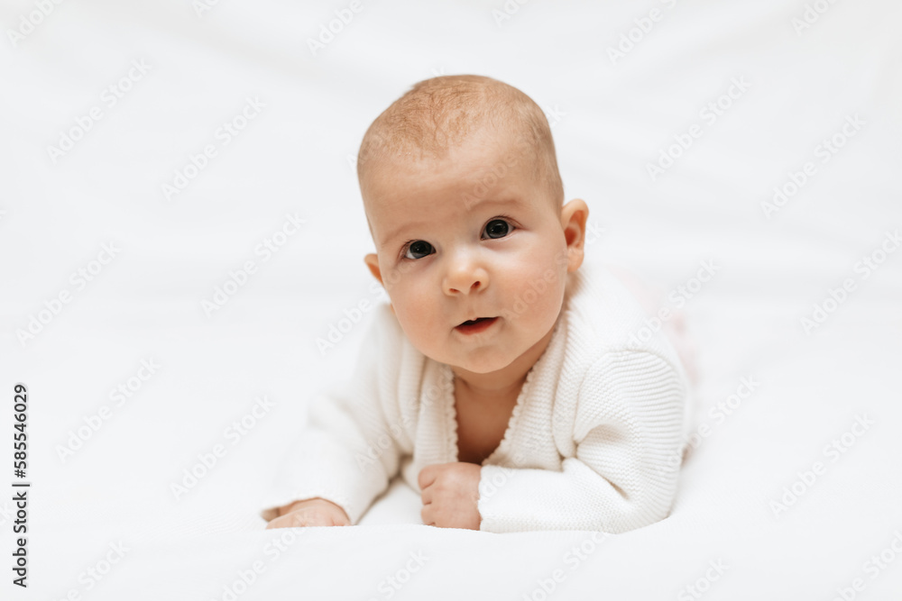 A newborn girl in a white knitted blouse is lying on her stomach leaning on her elbow on a white background. A newborn girl in a white knitted blouse is lying on her stomach leaning on her elbow on a white background.