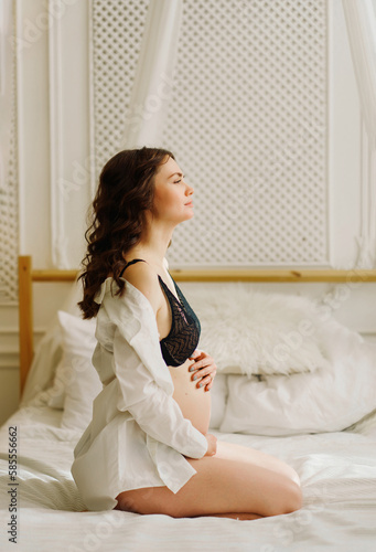 a slender pregnant woman in a black underwea and white shirt on the bed