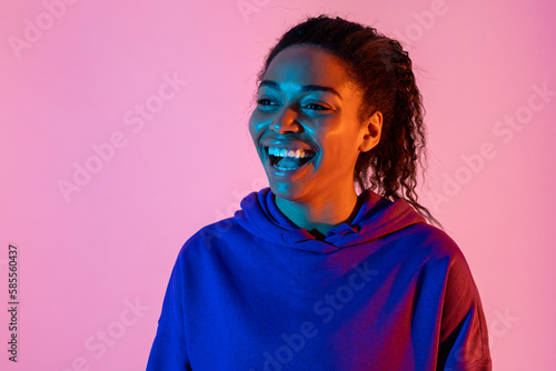 Portrait of happy young african american woman looking aside and smiling in pink neon light background, copy space