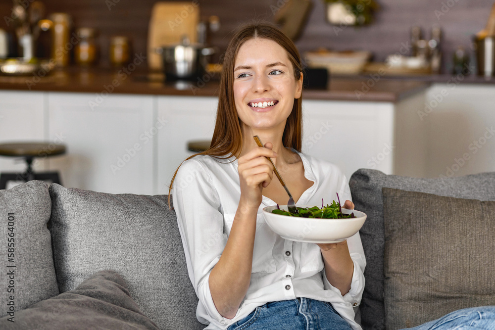 Smiling caucasian woman eating green salad sitting on sofa at home, healthy lifestyle, diet food concept