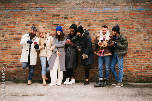 Mixed race group of stylish friends standing against a wall in outdoors smiling and using mobile phone.