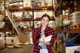 Young woman standing and checking availability order list during choosing items on shelf in storage warehouse