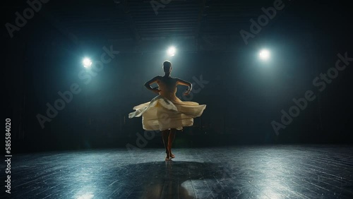 Graceful ballerina dancing elements of classical ballet on stage in backlight of spotlights. Silhouette of a young ballet dancer in pointe shoes and flowing dress