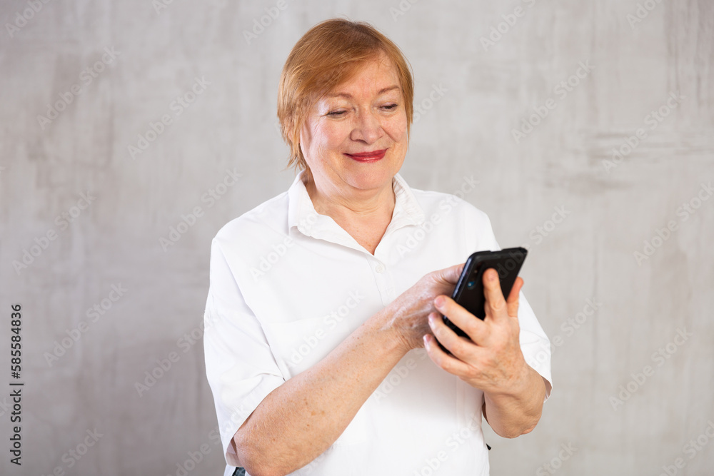 Fototapeta premium Smiling elderly woman in white shirt holding smartphone in hands and looking at screen with interest on gray background. Concept of tech-savvy and engaged with world around modern aged generation
