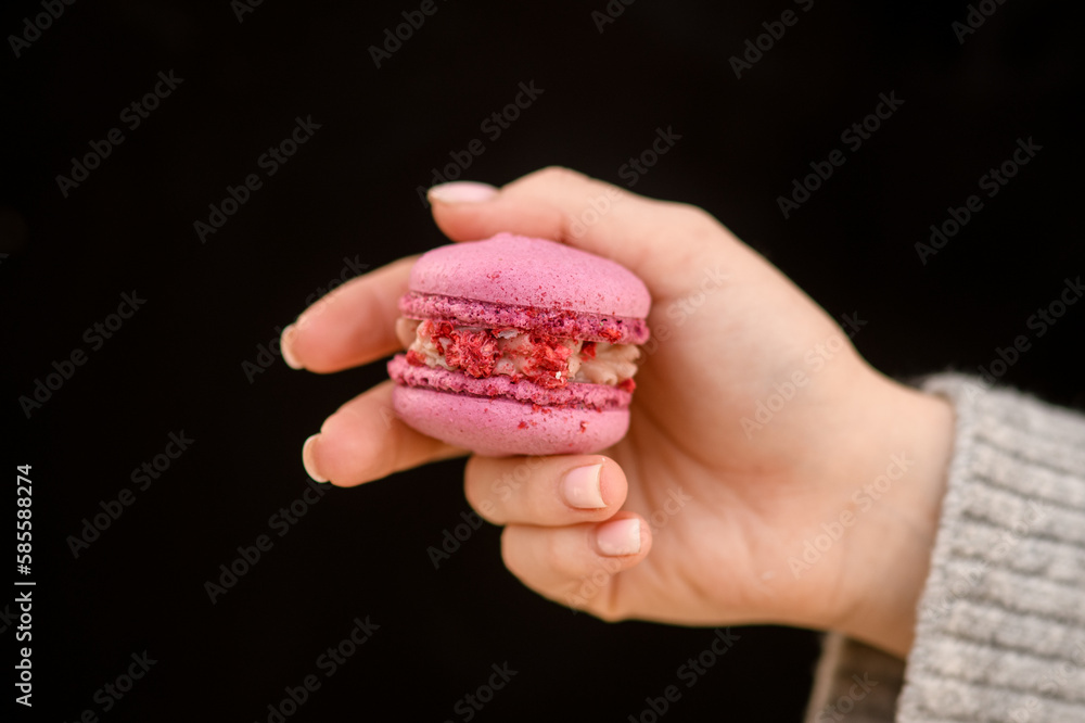Selective focus on sweet pink macaron in female hand on black background. Tasty dessert Selective focus on sweet pink macaron in female hand on black background. Tasty dessert