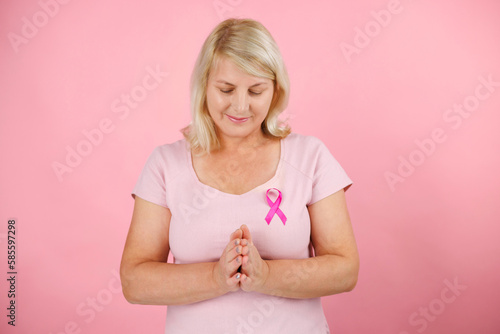 Mature woman praying on a pinck background. Wears a pink t-shirt and has a pink cancer ribbon, which means the fight against cancer. photo