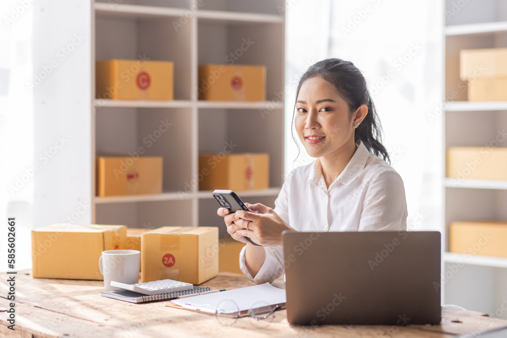 Asian SME business woman using laptop computer Phone checking customer order online shipping boxes at home. Starting Small business entrepreneur SME freelance. Online business, Work at home concept. Asian SME business woman using laptop computer Phone checking customer order online shipping boxes at home. Starting Small business entrepreneur SME freelance. Online business, Work at home concept.