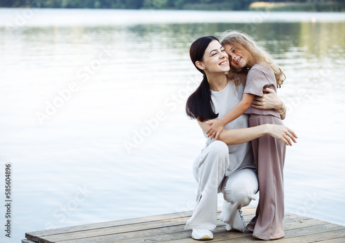 A young mother and her five-year-old daughter stand on the bridge by the lake and hug. Happy summer.