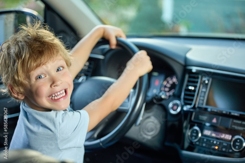 Happy boy is holding onto the steering wheel of the car, smiling broadly with joy. Smiling child in a modern car sits in the driver's seat, dreaming of winning a real race