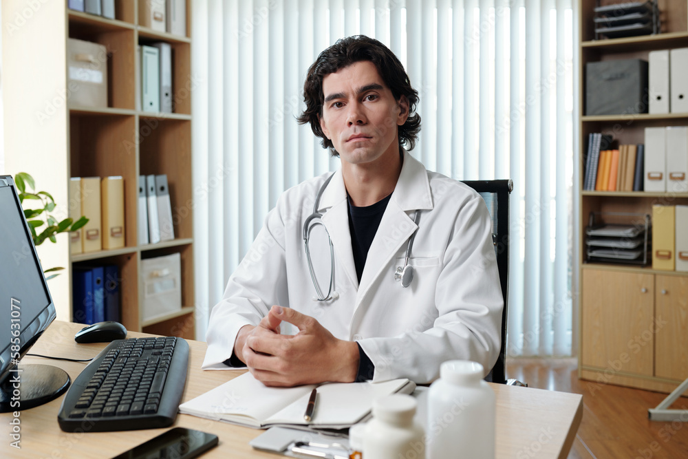 Young serious male teledoc in lab coat sitting by workplace in front of computer monitor in medical office and looking at camera Young serious male teledoc in lab coat sitting by workplace in front of computer monitor in medical office and looking at camera