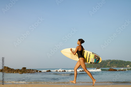 African american woman running with surfboard on ocean beach. Black female surfer with surf board. Pretty multiethnic girl goes on surfing session on tropical location at sunny sunrise.