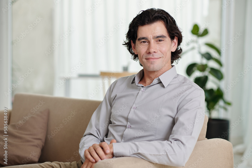 Young smiling businessman, blogger or psychologist in light blue shirt sitting on couch in home environment and looking at camera Young smiling businessman, blogger or psychologist in light blue shirt sitting on couch in home environment and looking at camera