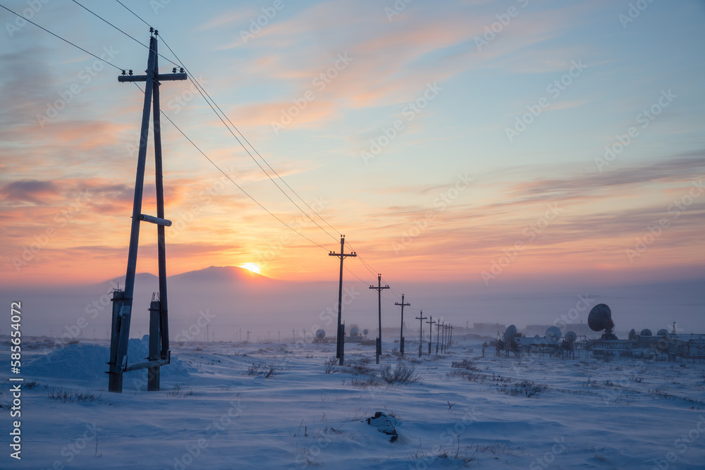 Electric poles and large satellite dishes in the snowy winter tundra. Winter arctic landscape. Cold frosty winter weather. The sun is setting behind the mountain. Chukotka, Siberia, Far East of Russia