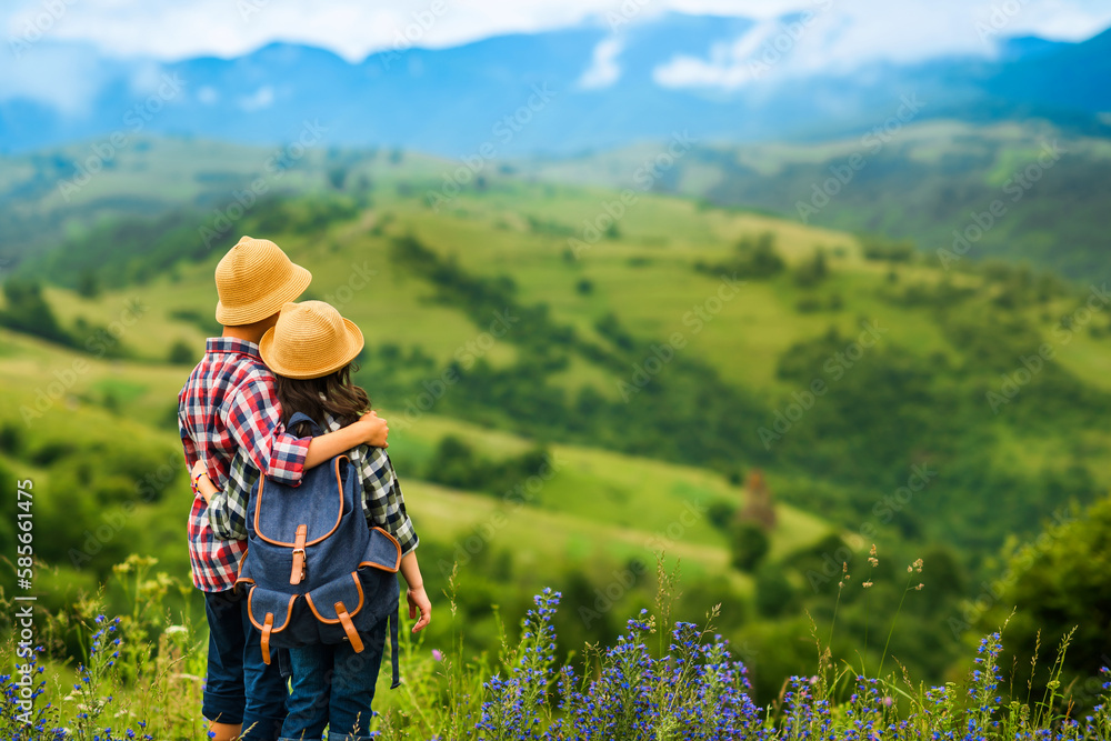 Two young children stand embracing and looking at the beautiful mountain landscape. Mountain hiking and tourism concept. Cloudy summer or spring day. Place for text to the right.