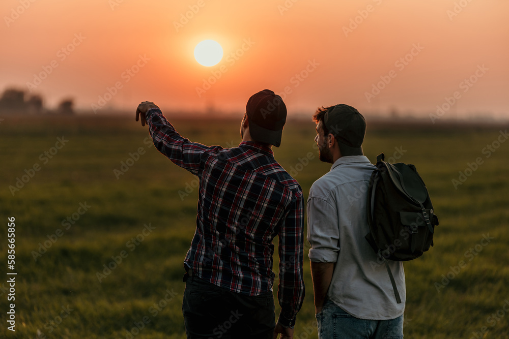 Rear view of a happy gay male couple tourist at the field at sunset outdoors during a hike in summer