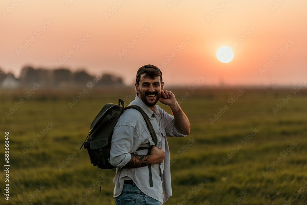 Fototapeta premium Portrait of a cheerful friendly male walking on the field with a backpack during a beautiful sunset outdoors.