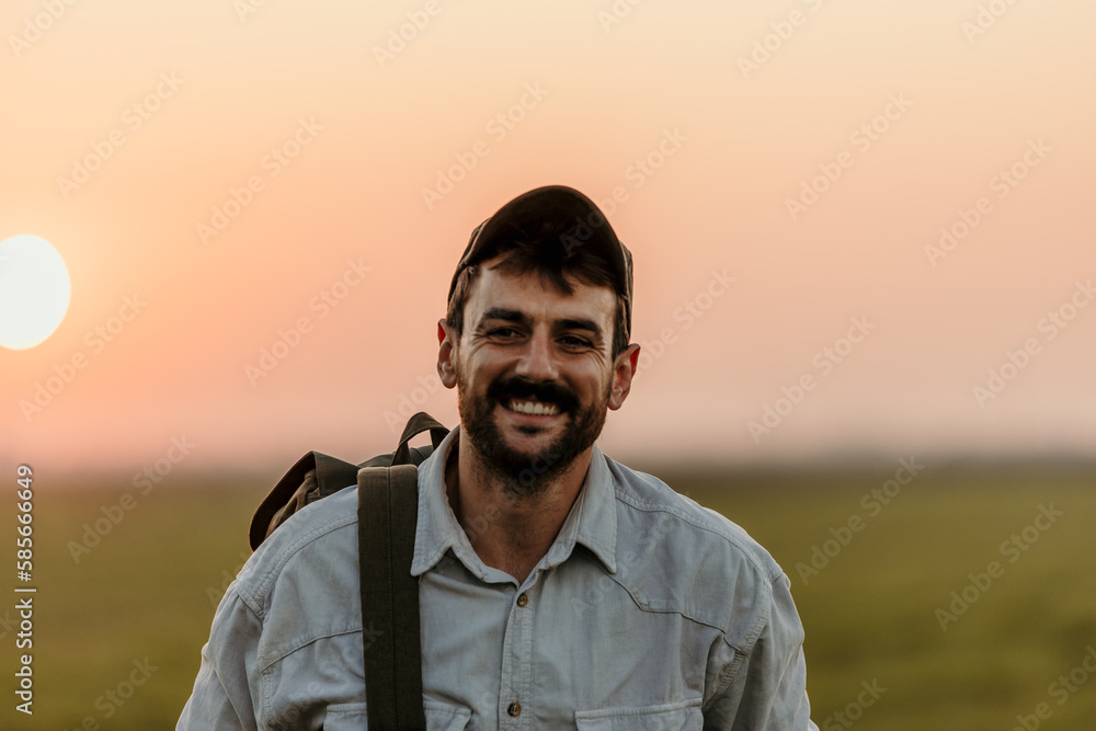 Portrait of a cheerful friendly male walking on the field with a backpack during a beautiful sunset outdoors.