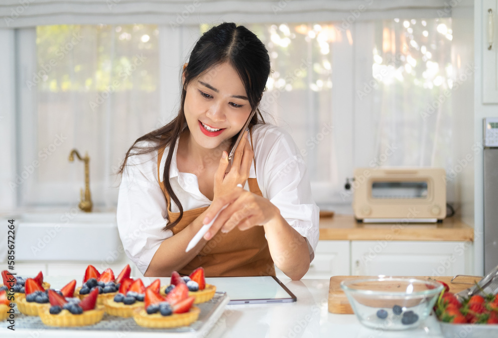 Portrait of happy asian female baker standing in own bakery, tapping on tablet device and speaking on smartphone. Female worker talking on mobile phone with client. Bakehouse concept Portrait of happy asian female baker standing in own bakery, tapping on tablet device and speaking on smartphone. Female worker talking on mobile phone with client. Bakehouse concept