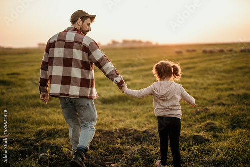 Rear view of a family on holiday vacation. Smiling father and little daughter holding hands and walking together on the meadow field. Parent with cute child girl relax and enjoy an outdoor lifestyle. © La Famiglia