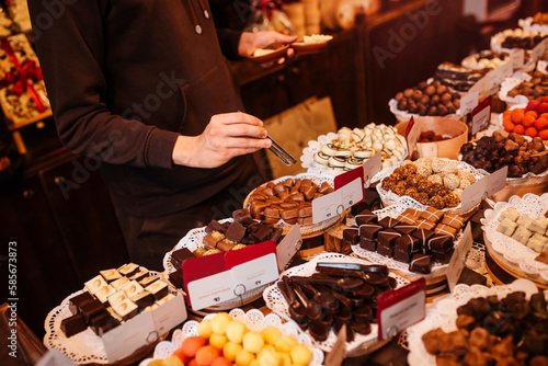 Chocolate workshop. Hands of seller of chocolates take candy with tweezers. Showcase with sweets.